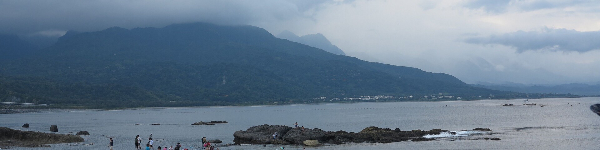 Una playa rocosa con aguas tranquilas, montañas lejanas y un cielo nublado.
