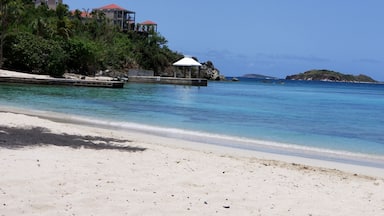 A beach with clear turquoise water, white sand, and a few boats. There are buildings with red roofs on a cliff in the background.