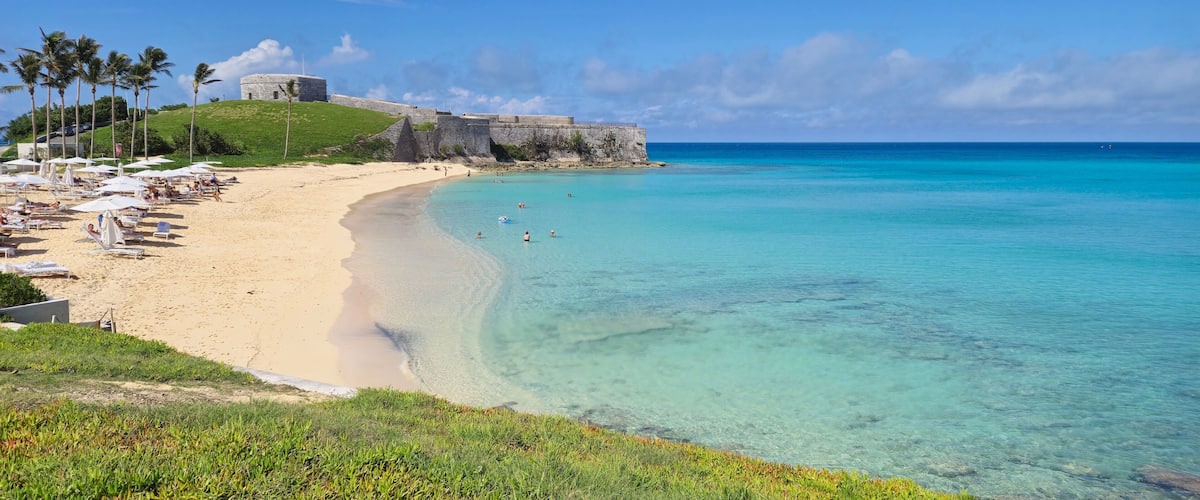 A beach with clear turquoise water, a sandy shore, and a historic fort on a grassy hill.
