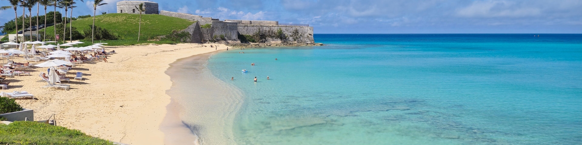 A beach with clear turquoise water, a sandy shore, and a historic fort on a grassy hill.