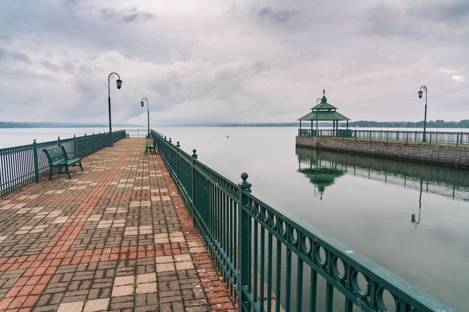 A brick-paved promenade with a green railing, a gazebo, and lampposts along a calm body of water.
