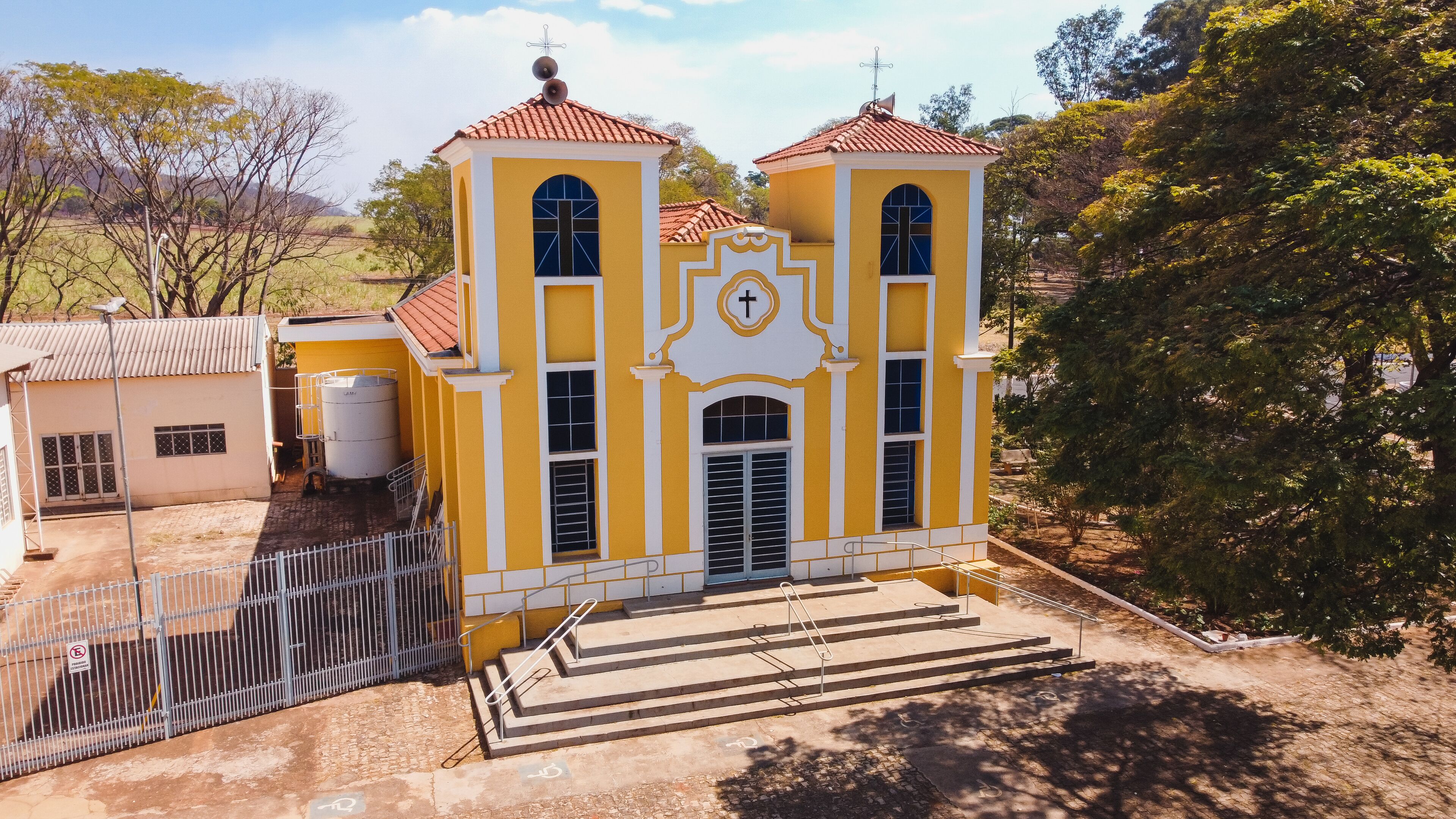 A yellow church with a cross on the facade, surrounded by trees and a fenced area.