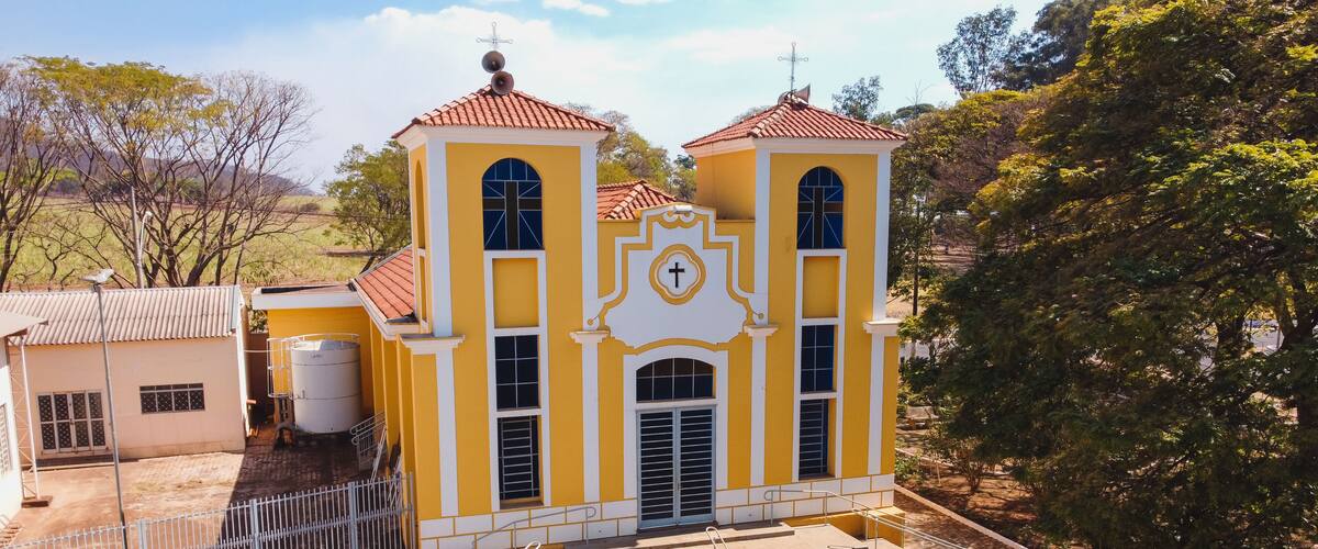 A yellow church with a cross on the facade, surrounded by trees and a fenced area.