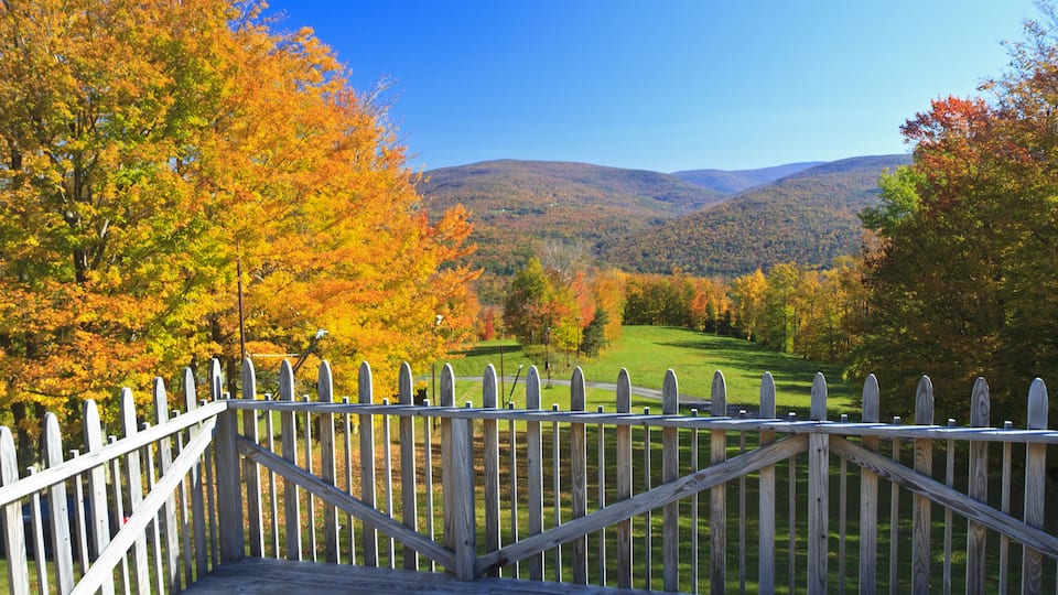 A wooden deck with a white picket fence overlooking a valley with autumn-colored trees and distant hills.