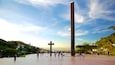 A large open plaza with a tall monument, people walking, and a scenic view of the city in the background.