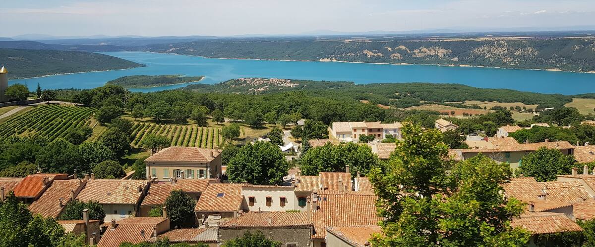 A coastal town with terracotta-roofed buildings, vineyards, and a large body of water.