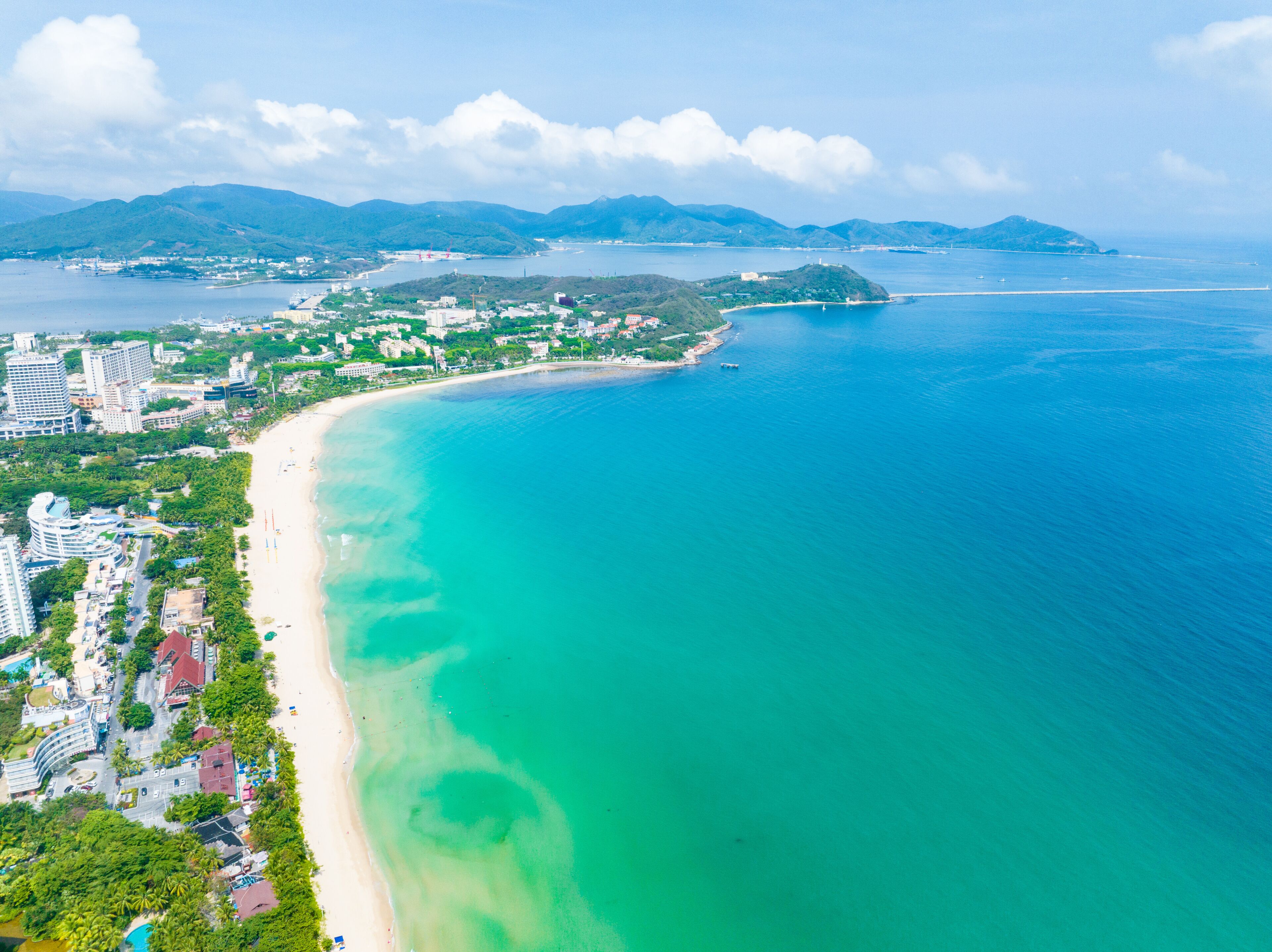 A coastal city with a long sandy beach, clear turquoise waters, and a backdrop of green hills.