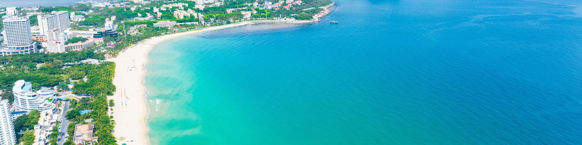 Uma cidade costeira com uma extensa praia de areia, águas turquesa cristalinas e colinas verdes ao fundo.