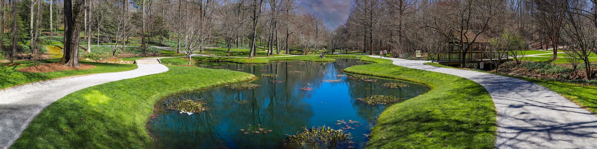 a panoramic shot of a still green pond surrounded by lush green grass, trees and plants with bare winter trees reflecting off the water with powerful red clouds at sunset at Gibbs Gardens