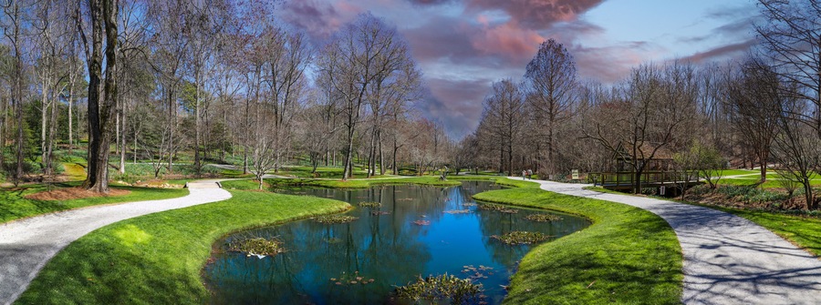 a panoramic shot of a still green pond surrounded by lush green grass, trees and plants with bare winter trees reflecting off the water with powerful red clouds at sunset at Gibbs Gardens