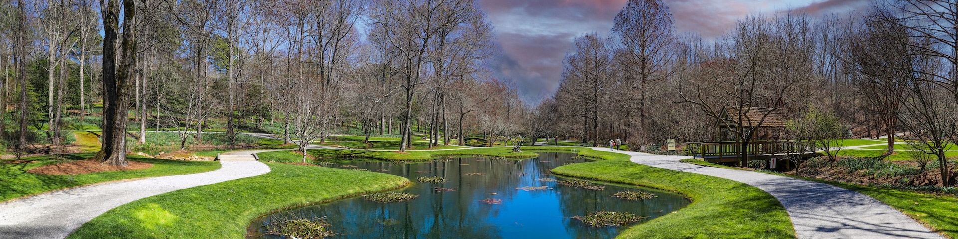 a panoramic shot of a still green pond surrounded by lush green grass, trees and plants with bare winter trees reflecting off the water with powerful red clouds at sunset at Gibbs Gardens