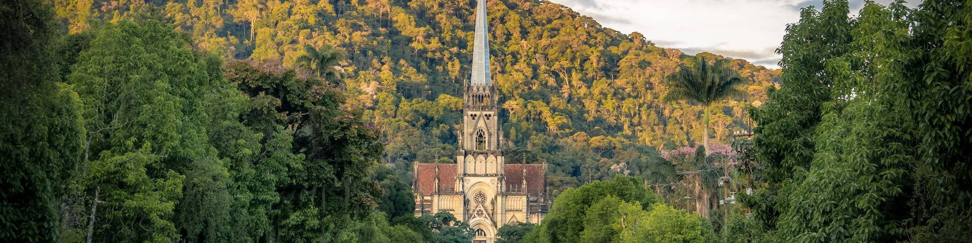 A cathedral with a tall spire, a river in the foreground, and a forested hill in the background.