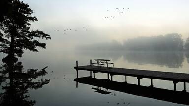 Un lago tranquilo con un muelle de madera y una mesa de picnic, rodeado de árboles y bruma.