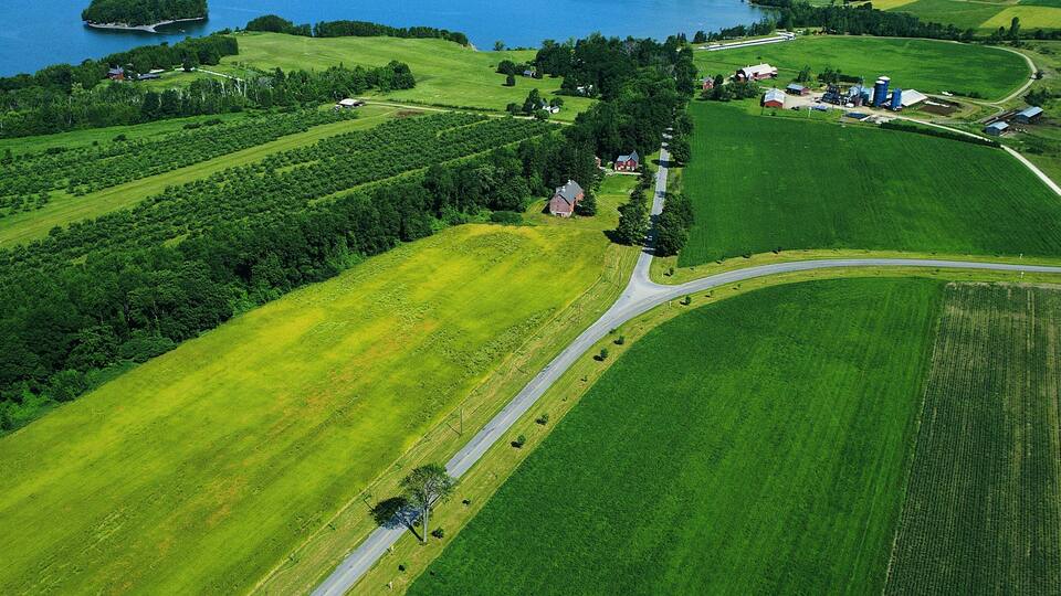 An aerial view of a coastal landscape with green fields, a winding road, and a small settlement with red-roofed buildings near the water.