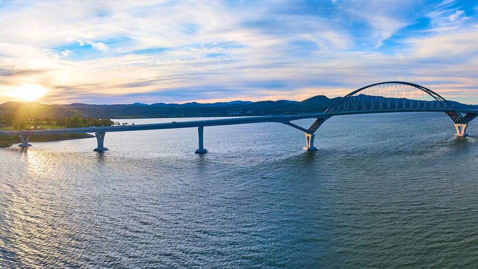 A bridge over a river with mountains in the background and the sun setting.