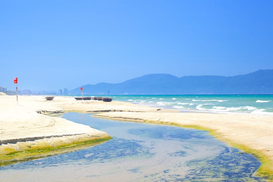 A beach with clear blue water, a small stream, and distant mountains.