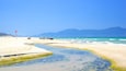 A beach with clear blue water, a small stream, and distant mountains.