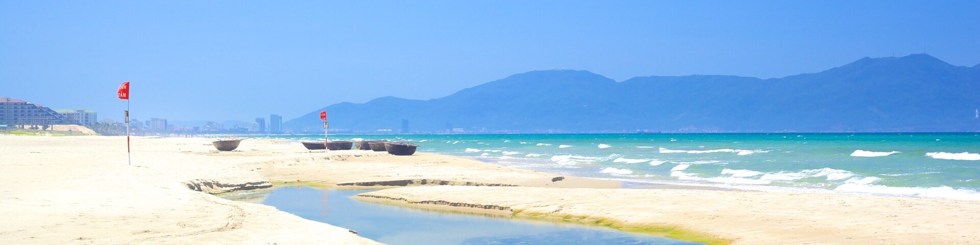 Una playa con agua azul cristalina, un pequeño arroyo y montañas a lo lejos.