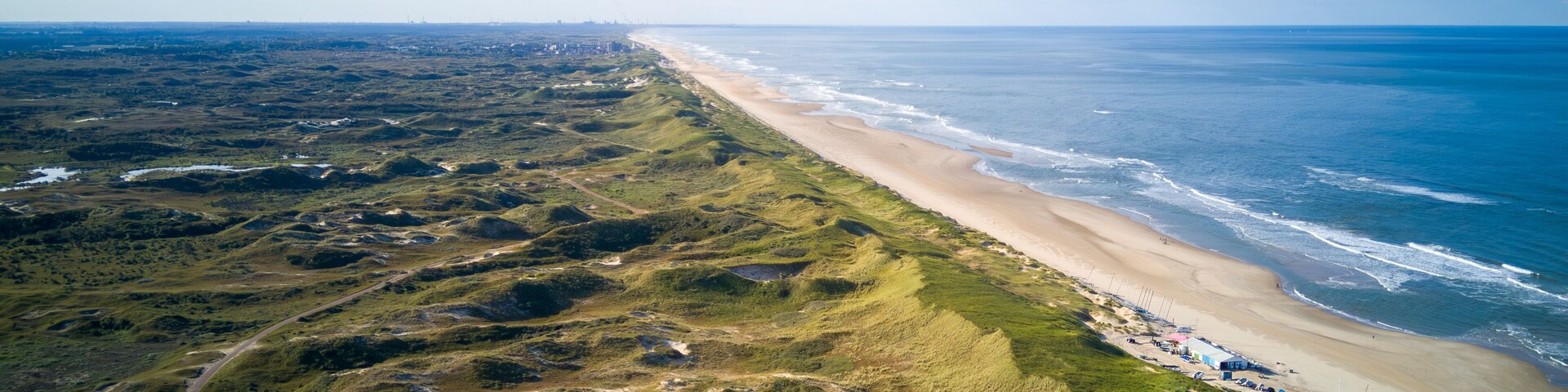 An aerial view of a coastal landscape with a sandy beach, grassy dunes, and a clear blue sky.