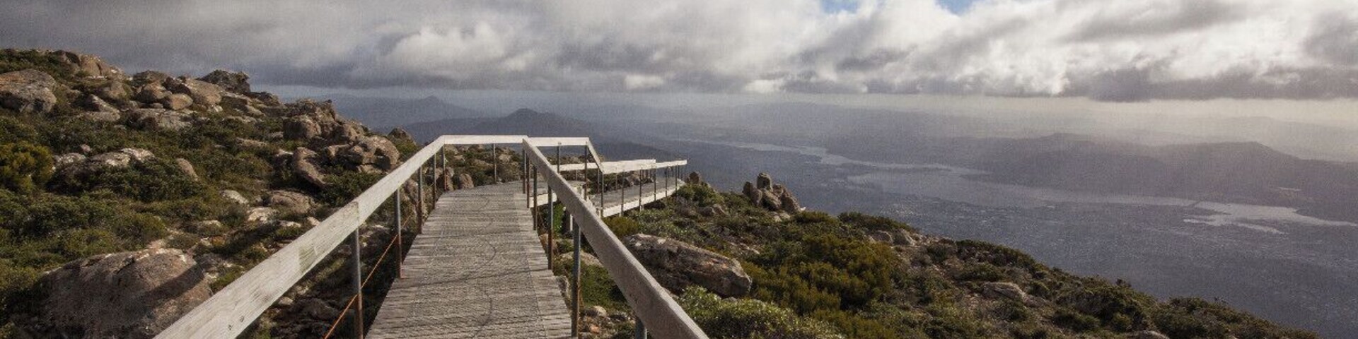 Pasarela de madera que conduce a un mirador con vistas a un paisaje costero de terreno rocoso y una vista distante del mar.
