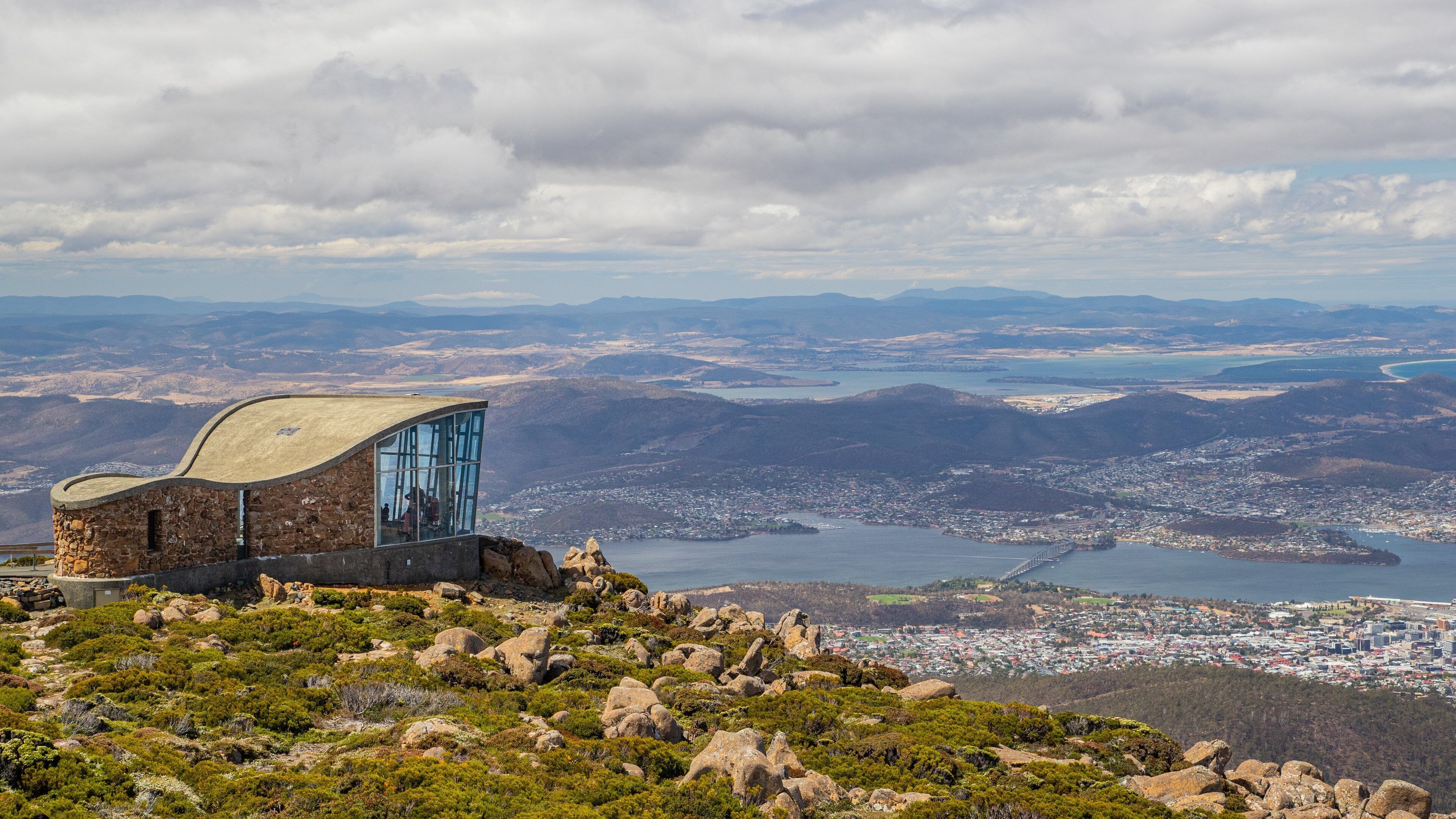 A stone building with a glass facade overlooking a coastal city and mountains.