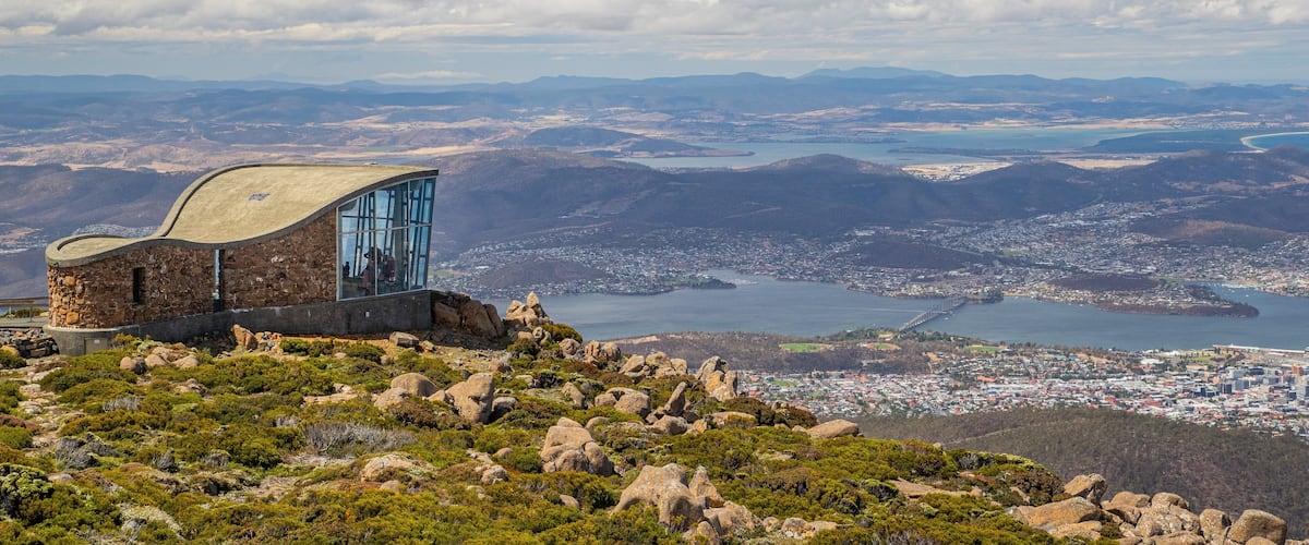A stone building with a glass facade overlooking a coastal city and mountains.