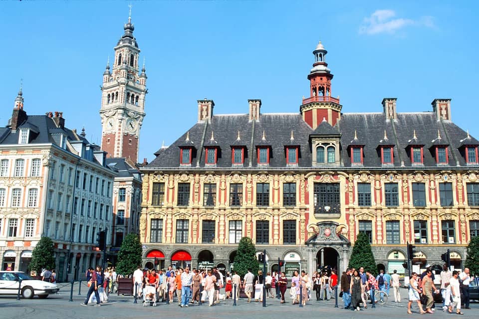 Un bâtiment historique à l’architecture riche et élaborée, doté d’un imposant beffroi et donnant sur une place animée où les piétons se promènent.