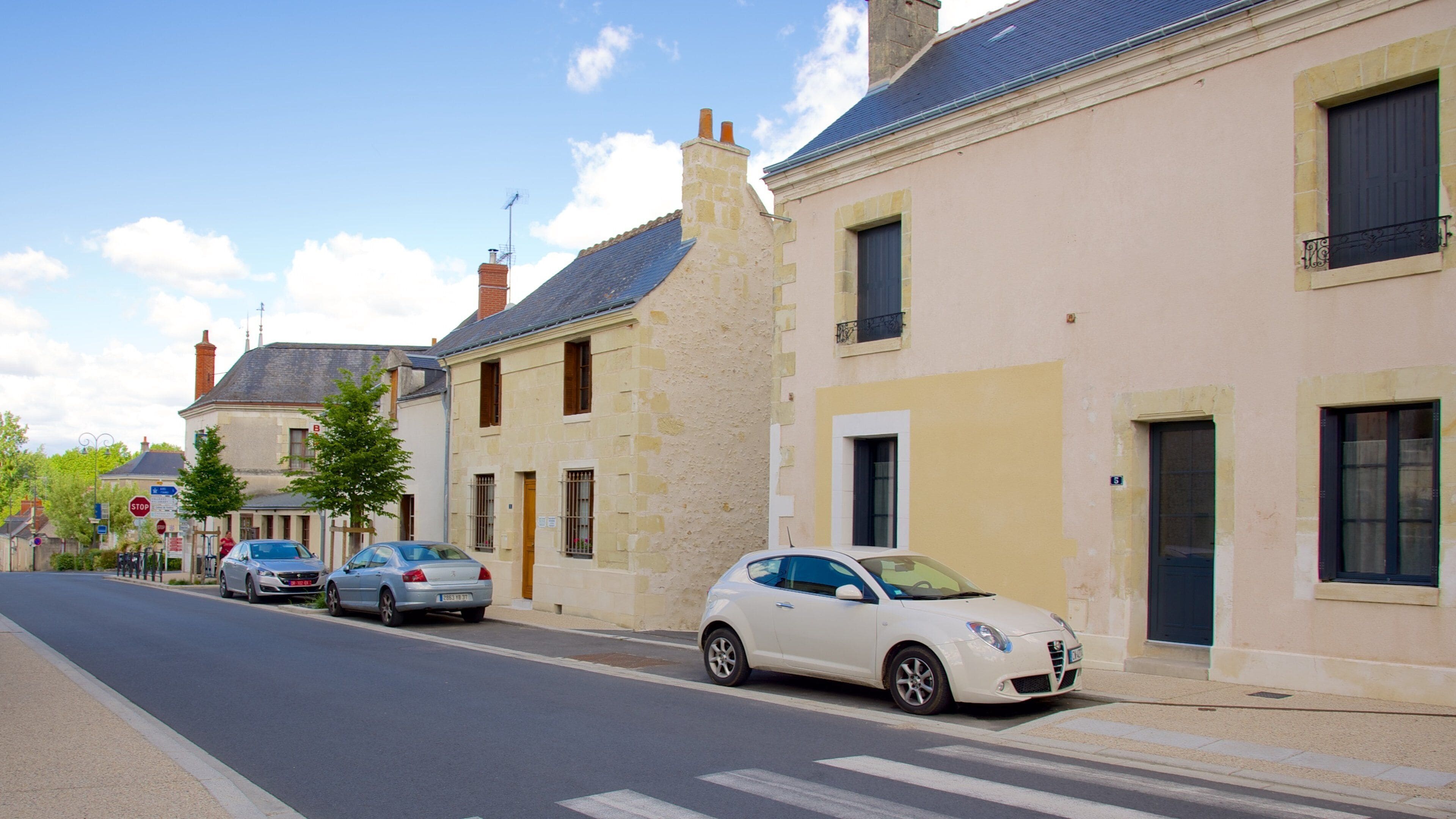 Una calle con coches estacionados, una señal de parada y edificios con fachadas de piedra y techos azules.