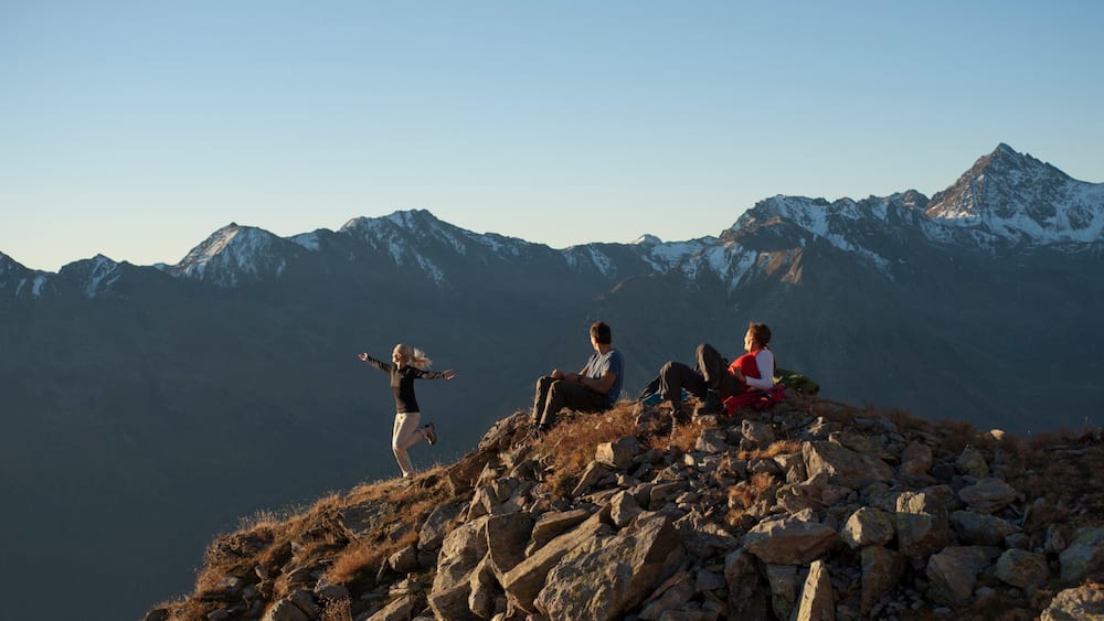 Trois personnes se tiennent au sommet d’une montagne rocheuse, avec des montagnes enneigées en arrière-plan.