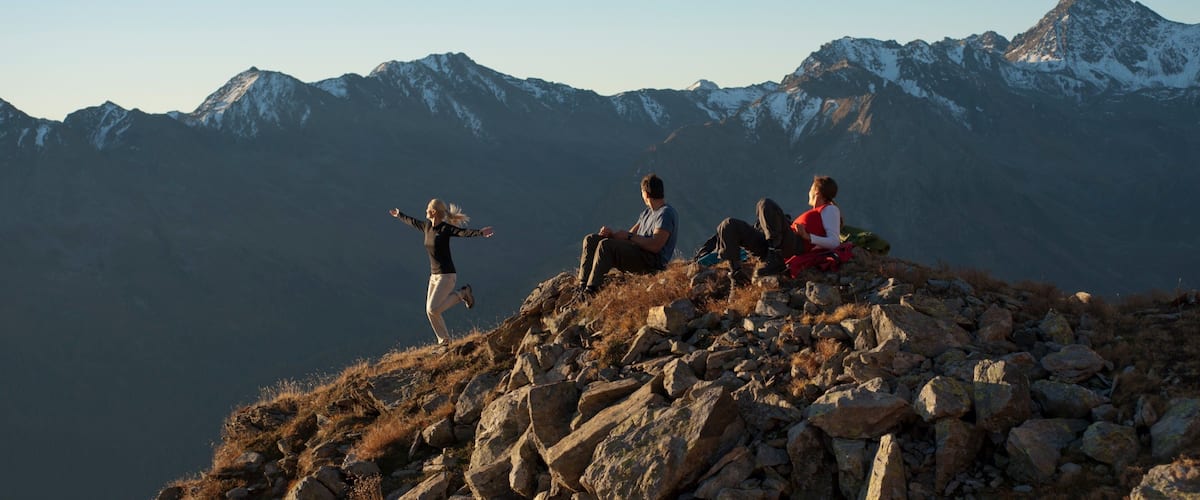 Tres personas en la cima de una montaña rocosa, con montañas nevadas al fondo.