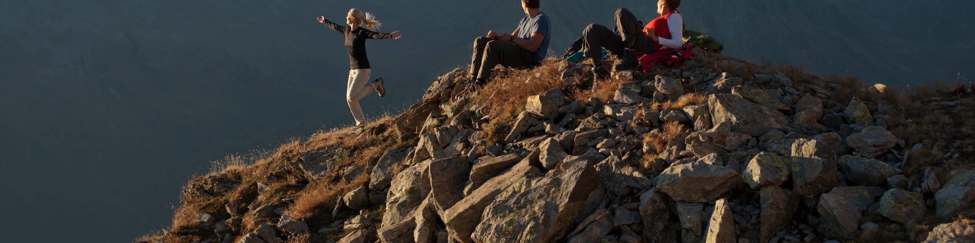Three people on a rocky mountain peak with snow-capped mountains in the background.