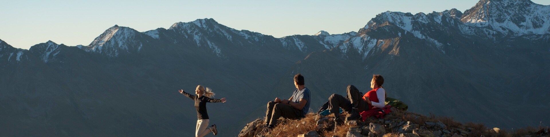 Três pessoas no cume de uma montanha rochosa, com montanhas nevadas ao fundo.