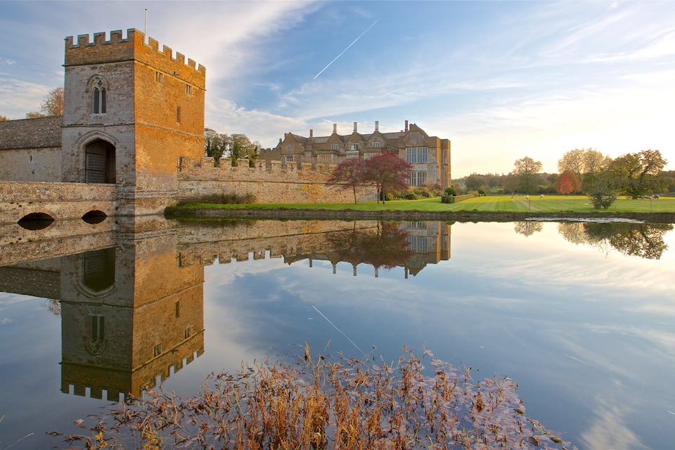 A historic castle with a tower and a large manor house across a reflective lake.