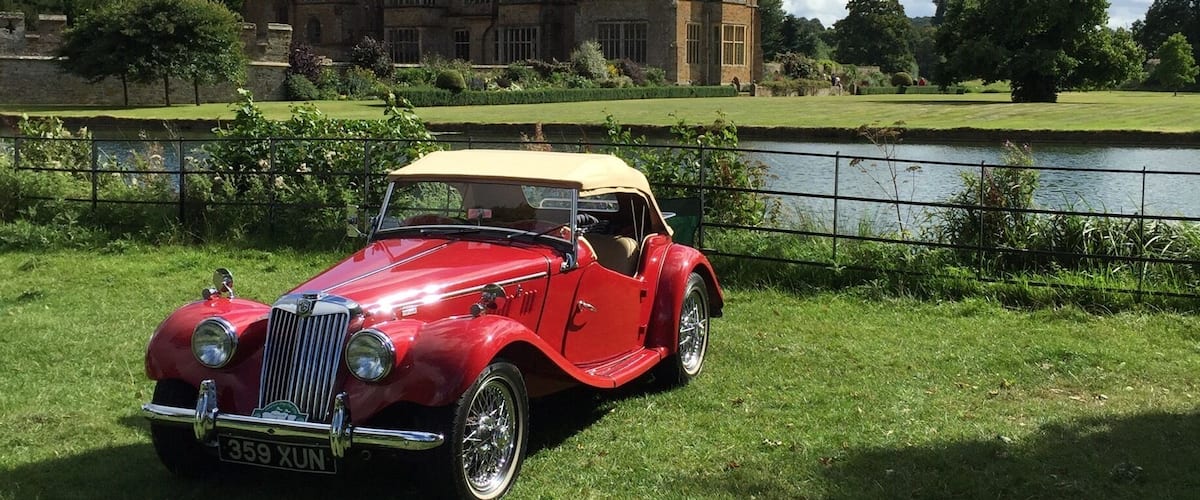 A red vintage car parked on grass with a large building in the background.