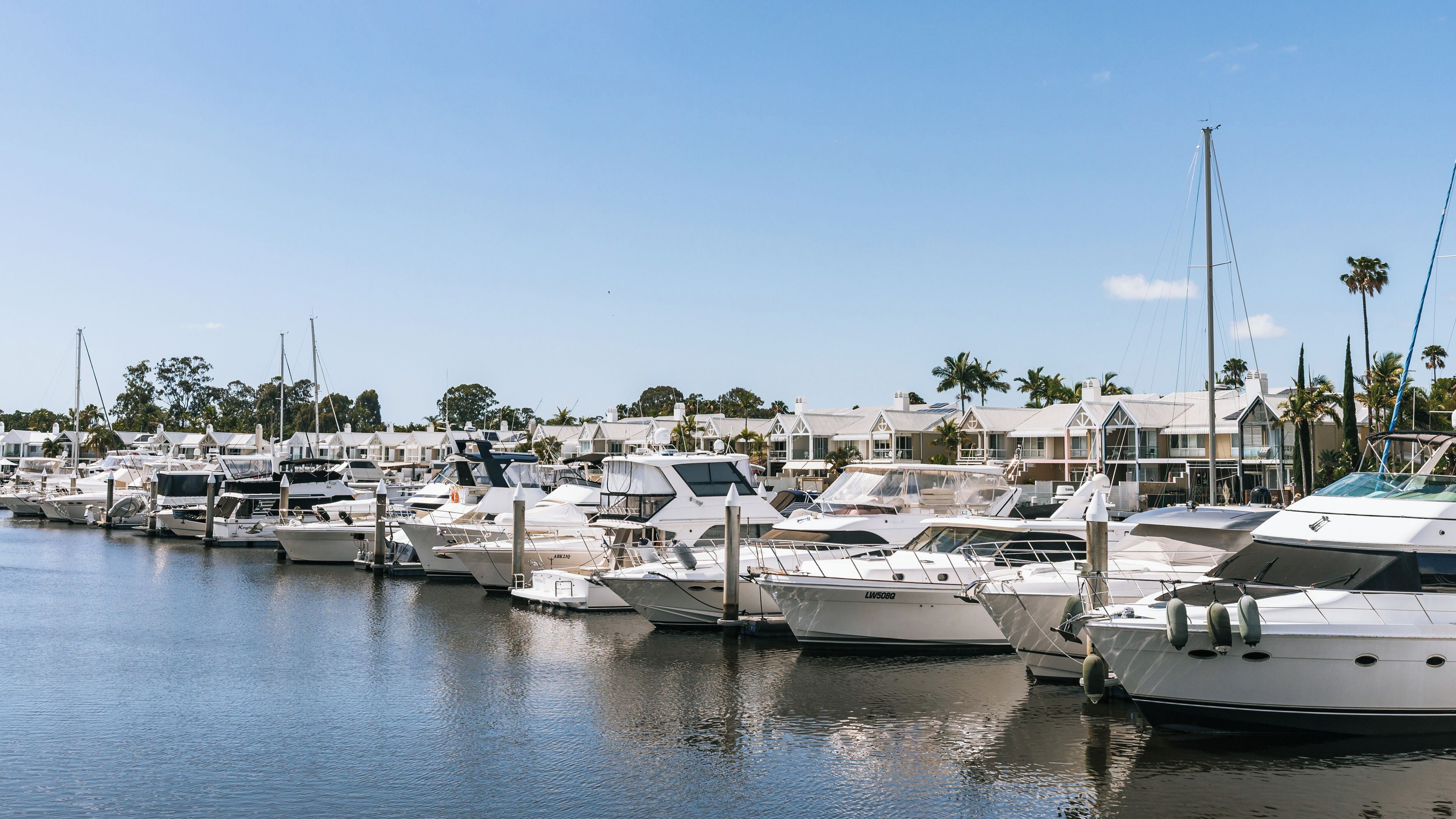A marina with multiple yachts docked, surrounded by palm trees and houses.