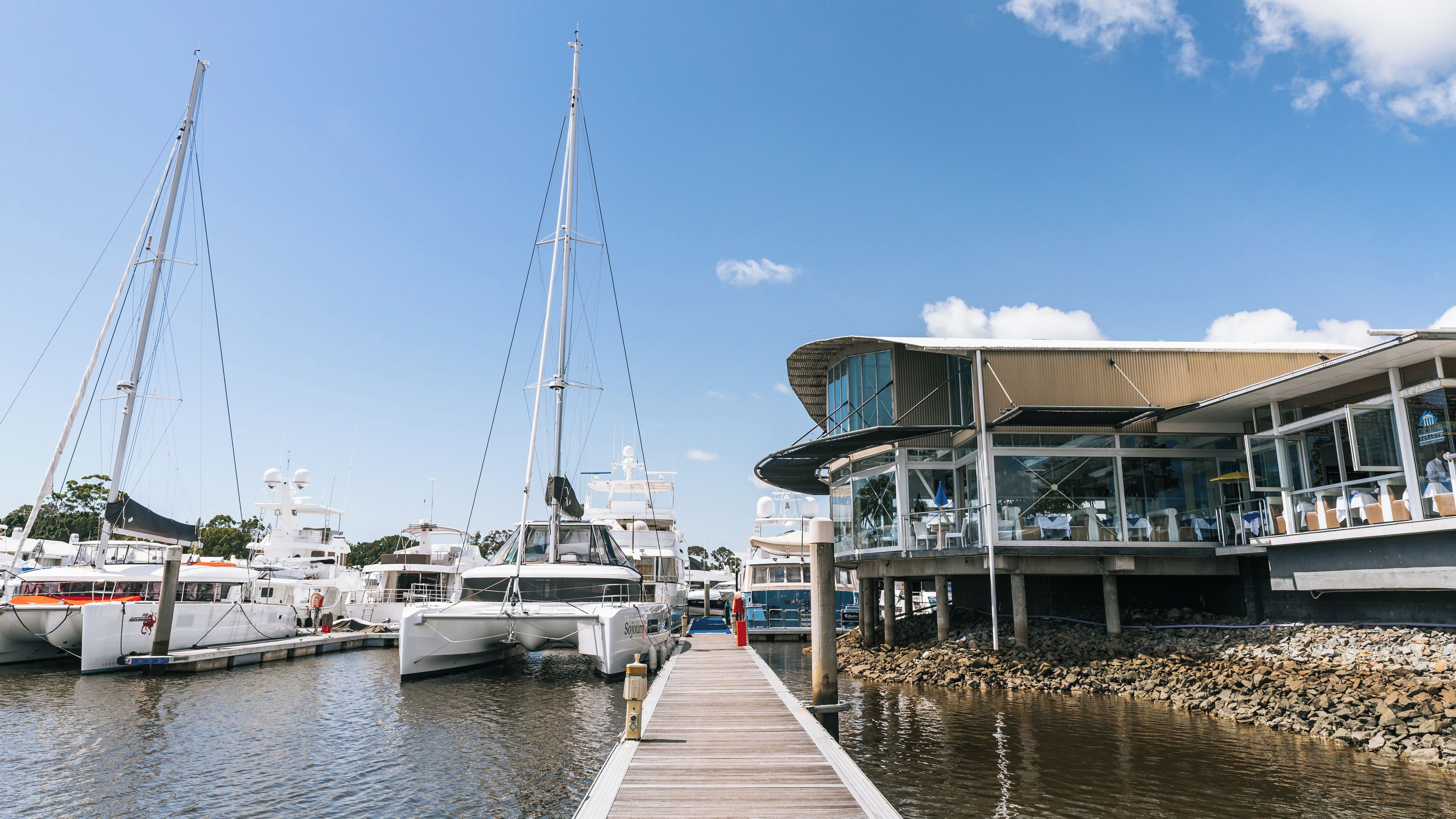 A marina with several sailboats docked, a wooden pier, and a modern building with outdoor seating.