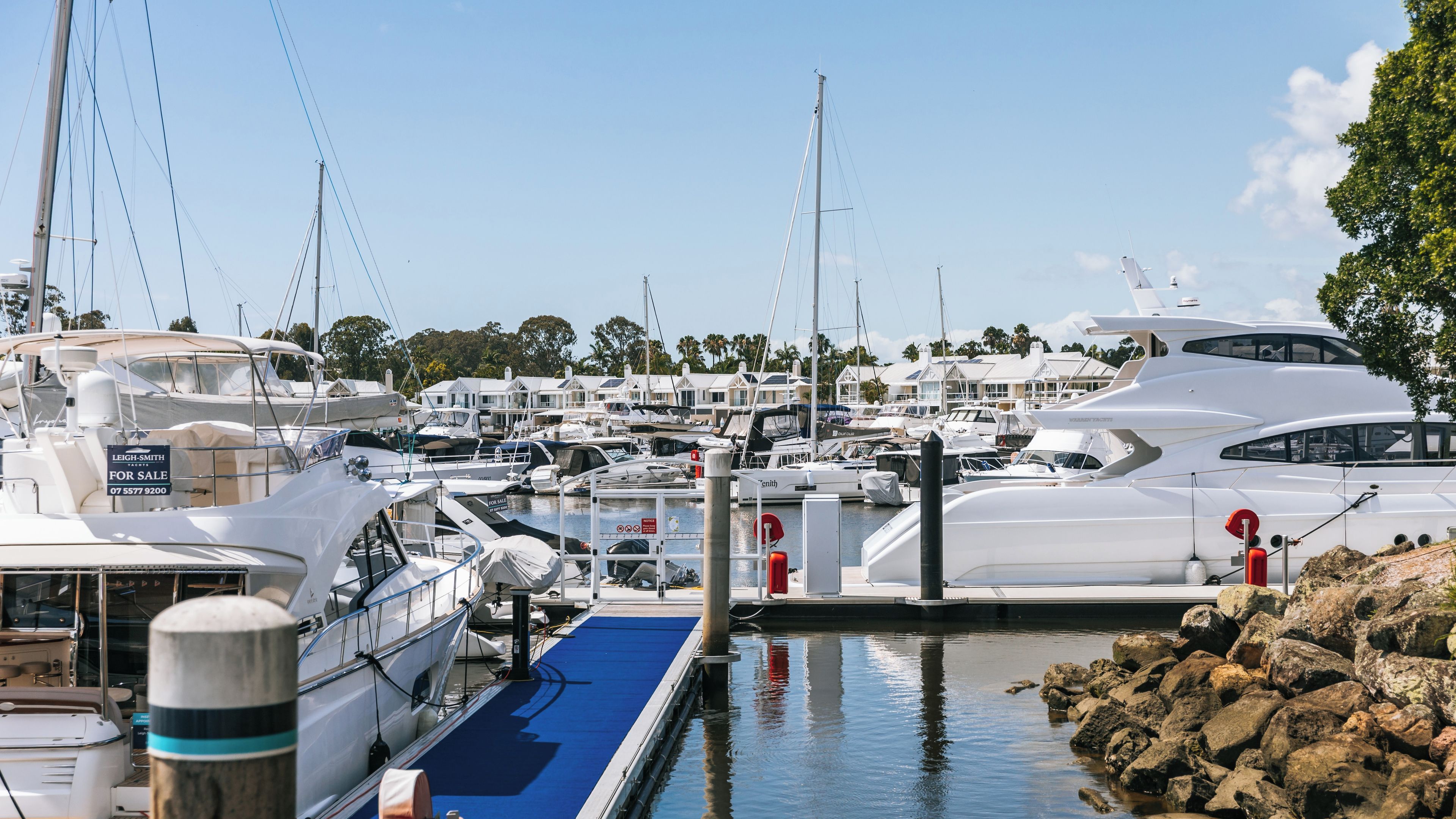 A marina with several yachts docked, a blue slipway, and a row of houses in the background.