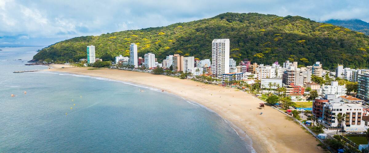 A coastal city with a sandy beach, tall buildings, and a forested hill in the background.