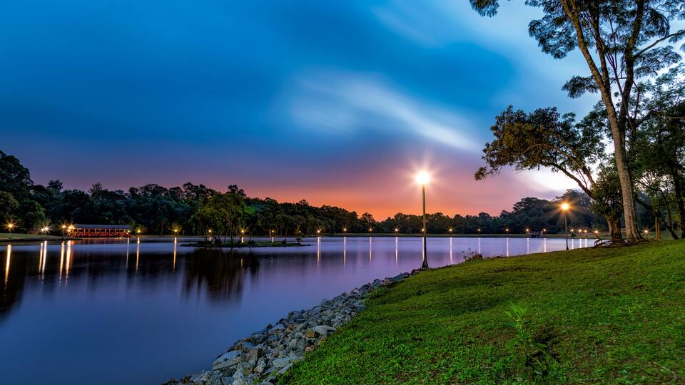 A tranquil lakeside scene at dusk with a bridge, trees, and a well-lit path.