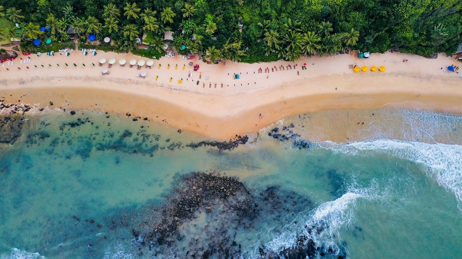 Vue aérienne d’une plage aux eaux turquoise et limpides, bordée d’un rivage sablonneux, avec une forêt dense en arrière-plan.