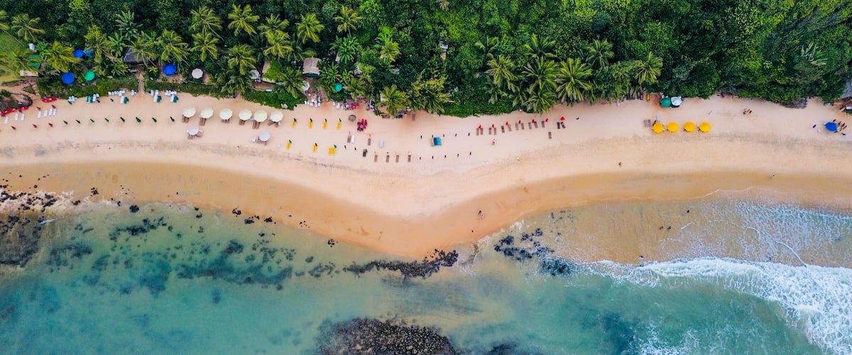 An aerial view of a beach with clear turquoise waters, a sandy shore, and a dense forest in the background.