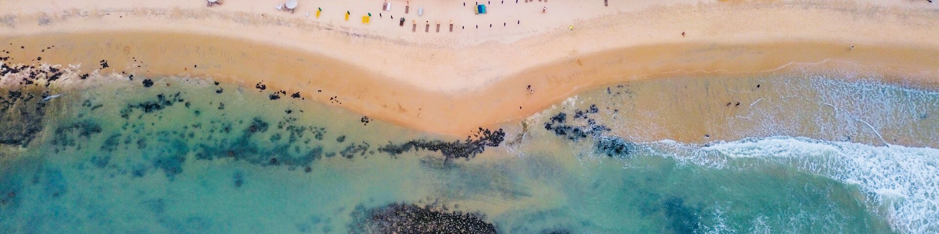 An aerial view of a beach with clear turquoise waters, a sandy shore, and a dense forest in the background.
