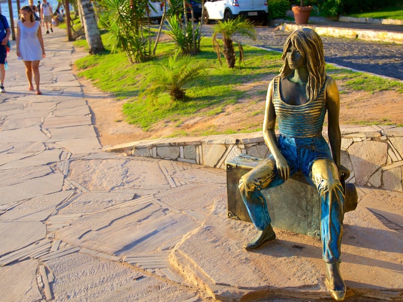 A bronze statue of a woman sitting on a stone bench by a waterfront.