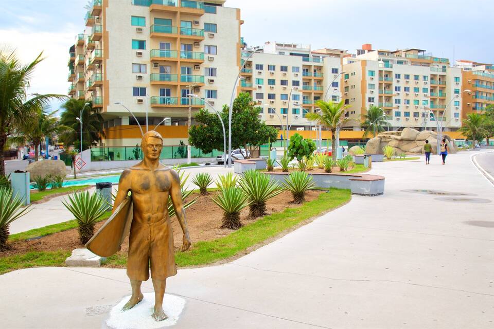 Estátua de bronze de um homem com capa, numa calçada, rodeada por palmeiras e um edifício moderno ao fundo.