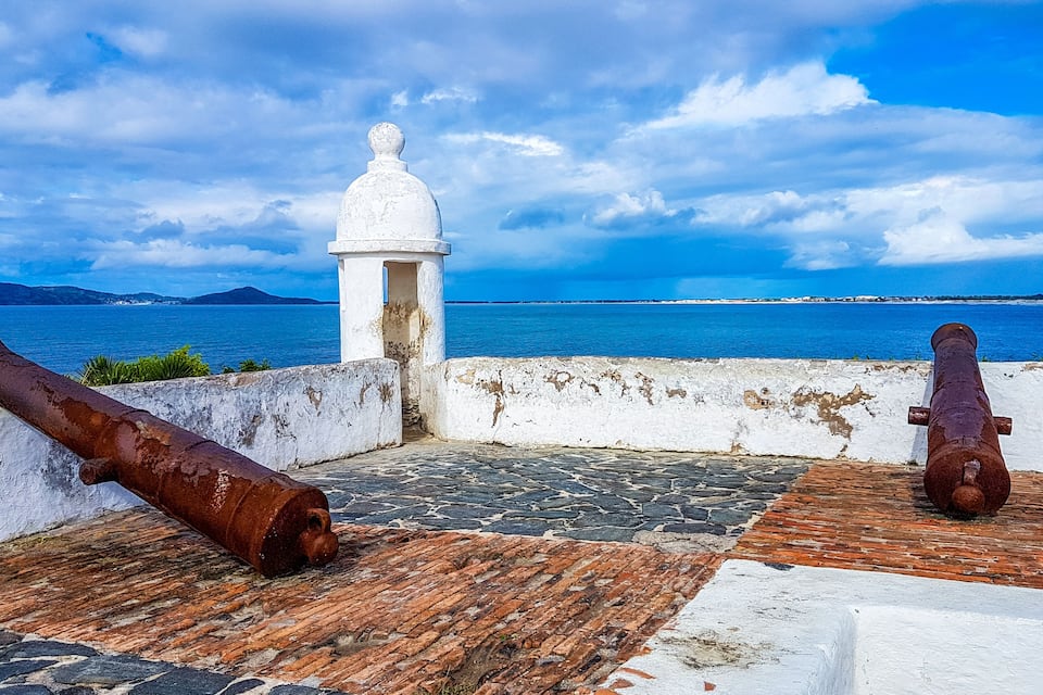 A coastal fort with cannons, a white tower, and a view of the sea and distant mountains.