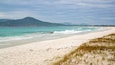 A beach with clear turquoise water, white sand, and a distant hill with a tower.
