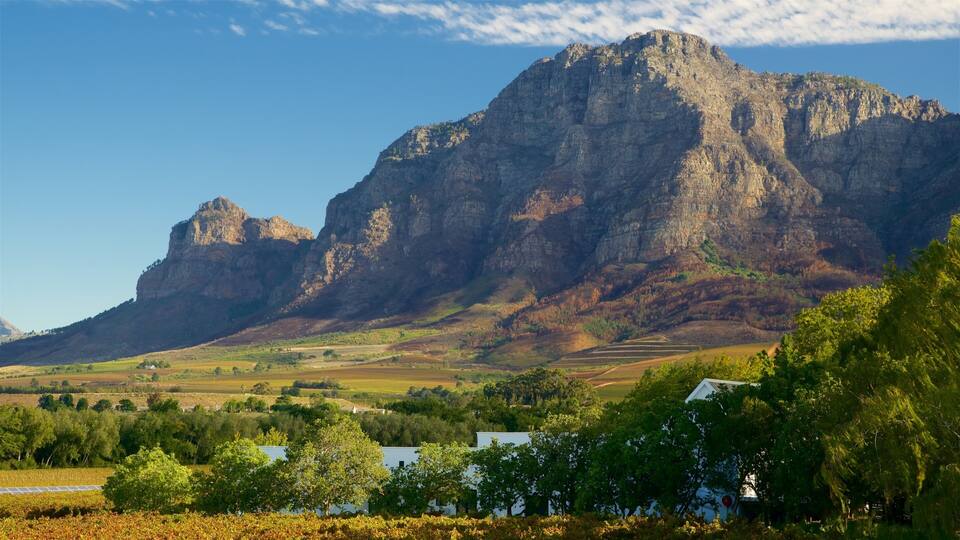 A mountain range with a prominent peak, a valley with fields, and a small white building in the foreground.