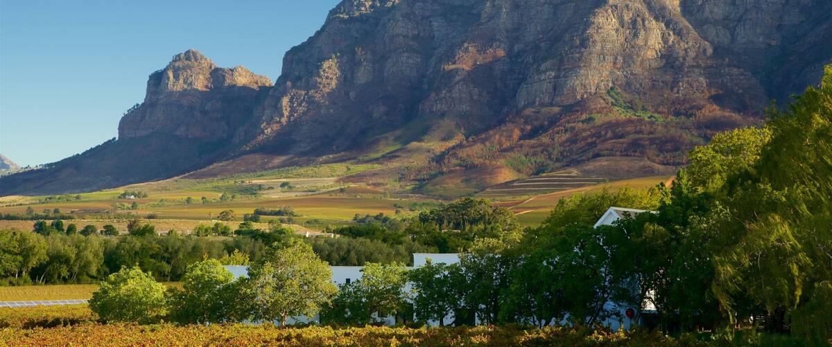 A mountain range with a prominent peak, a valley with fields, and a small white building in the foreground.