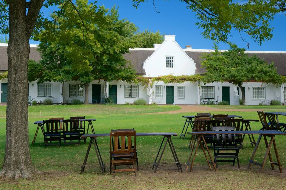 Un edificio de estilo colonial blanco con contraventanas verdes, rodeado de un césped con sillas y mesas de madera.