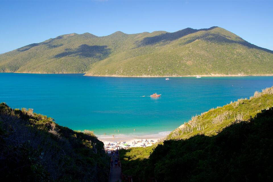 A coastal landscape with a beach, clear turquoise waters, and green hills in the background.